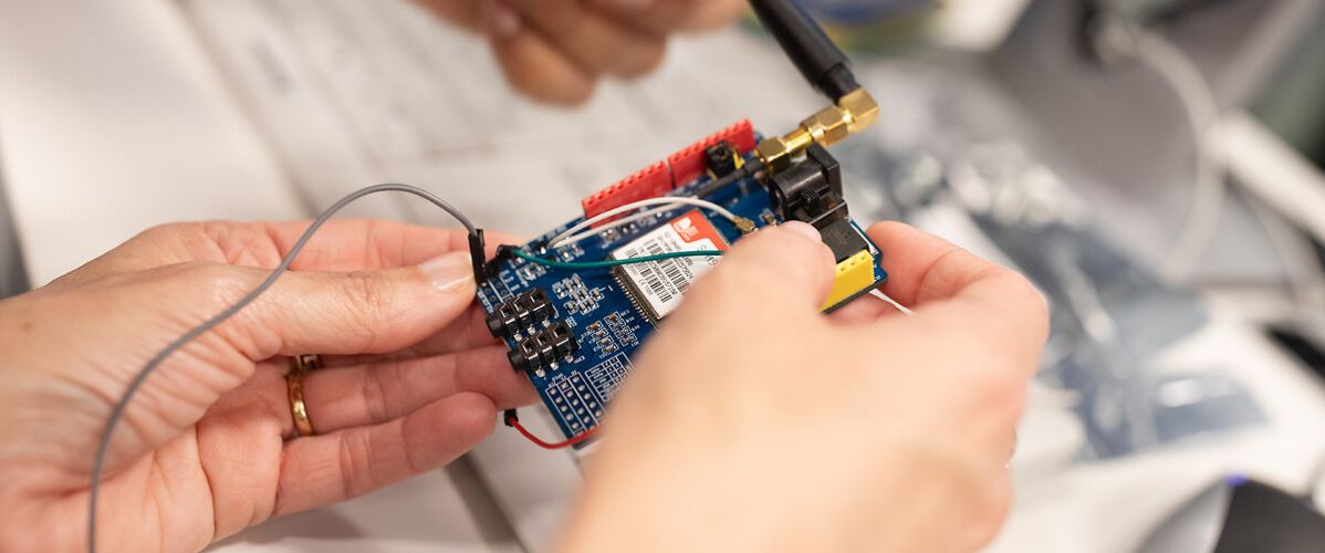Alt text: Hands are handling an electronic circuit board fitted with components, wires, and an antenna. The person is connecting or adjusting parts of the device on a technical work surface.