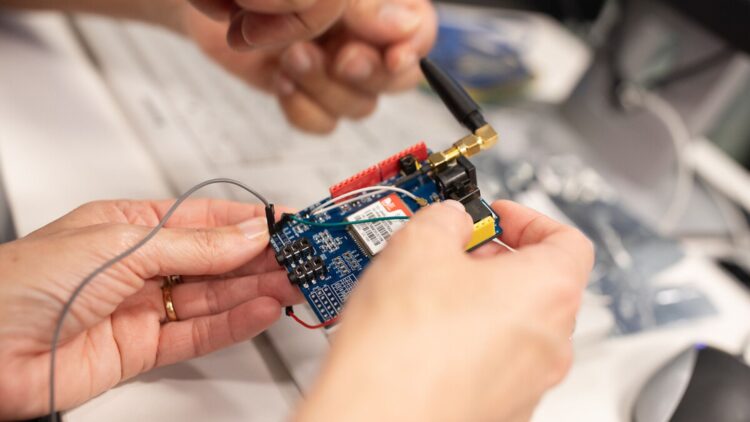 Des mains manipulent une carte électronique équipée de composants, de câbles et d’une antenne. La personne est en train de connecter ou d’ajuster les éléments du circuit, posé sur un plan de travail technique.